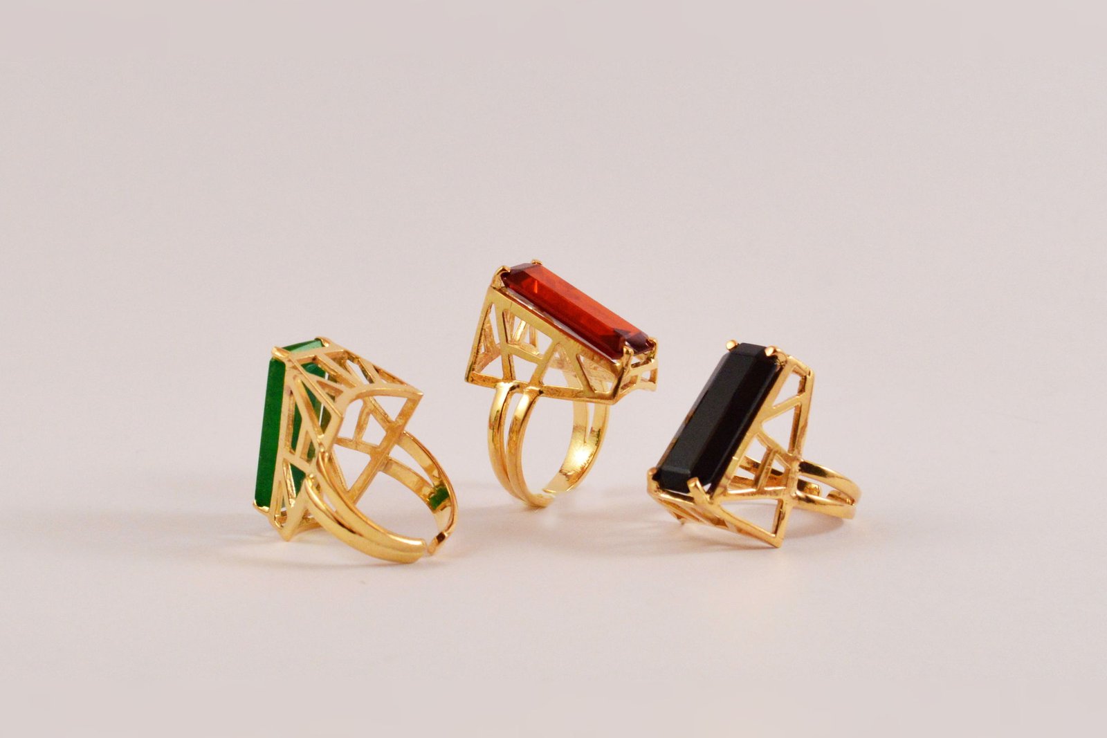Three Statement Rings with open architectural cages set with jade, red zircon and black onyx, on a pale blush ground.