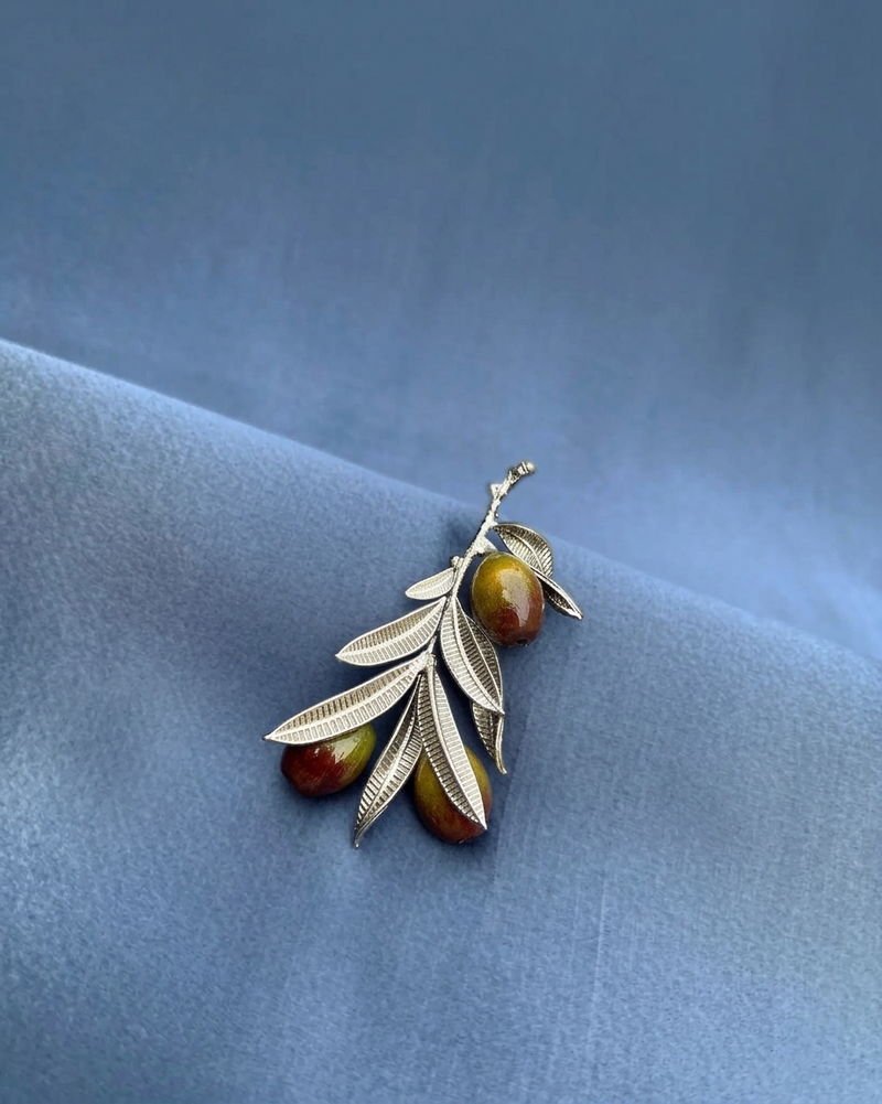 The Olive Branch brooch in silver with hand-painted olives, resting on folded blue cloth.