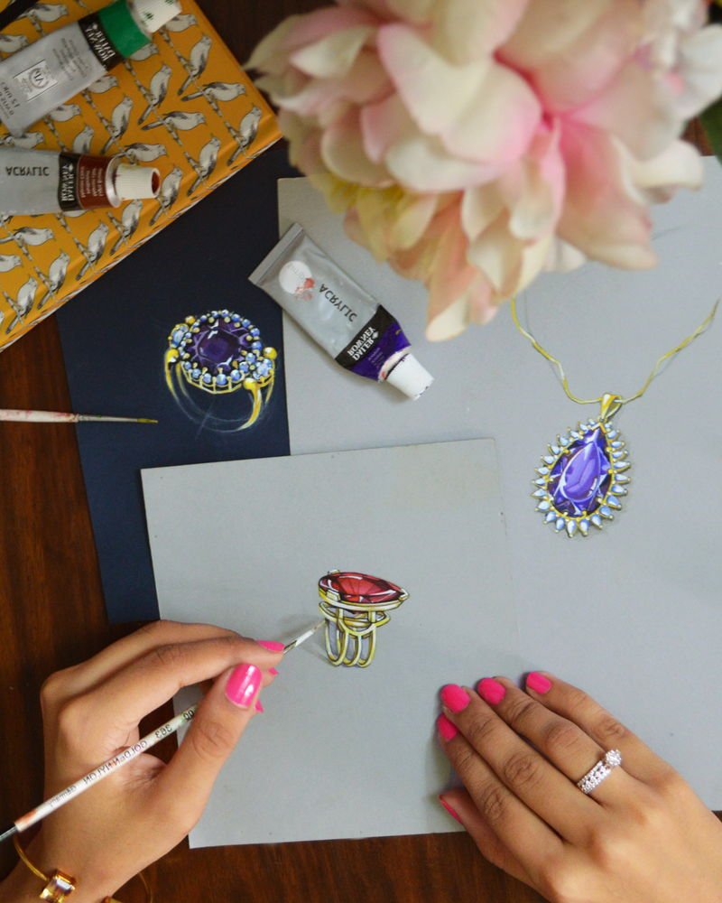 Two hands painting a gouache rendering of a ring on grey paper, beside paint tubes and a rendering of a pendant.